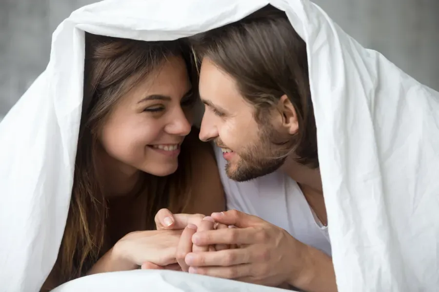 Couple holding hands lovingly under white bed sheets